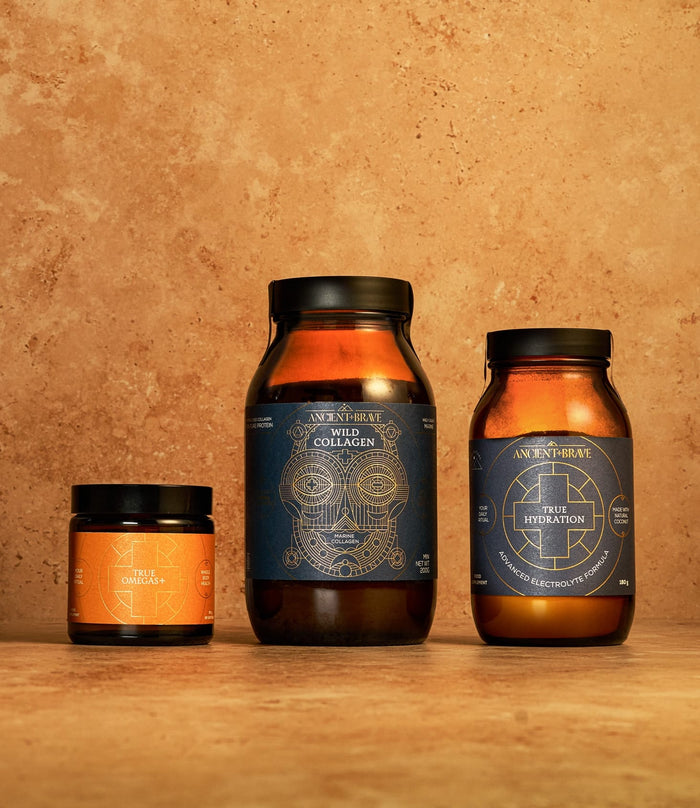 Three amber jars of the Ancient + Brave Beauty Kit, featuring blue and orange labels, arranged on a textured tan surface against a matching tan background—formulated to support hair growth and skin elasticity.