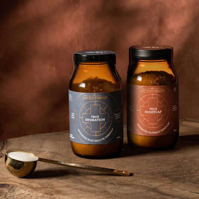 Two jars labeled Dawn To Dusk Duo from Ancient + Brave sit on a wooden surface, with a gold spoonful of powder beside them—perfect additions to your daily rehydration routine—against a warm brown background.