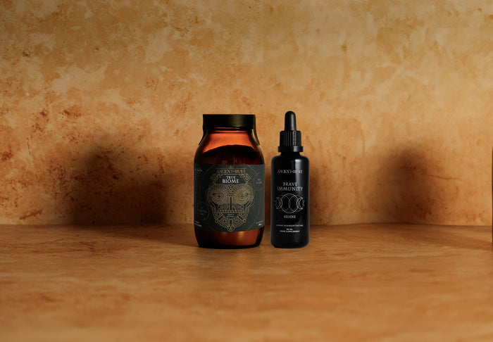 Two dark-colored jars from Ancient + Brave’s Inner Strength Ritual sit on a marble surface against a beige, textured background. The larger jar, topped with a metallic lid, and the smaller dropper bottle both display intricately designed labels—reflecting formulas thoughtfully crafted for gut health and immune support.