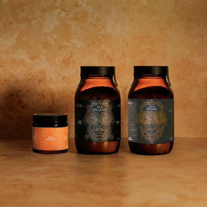 Three amber-colored supplement jars from Ancient + Brave, each with detailed geometric labels, are arranged on a tan surface. Designed to support gut health and immune balance as part of The Gut Health Kit, one small jar sits to the left of two larger jars—each featuring unique product names.