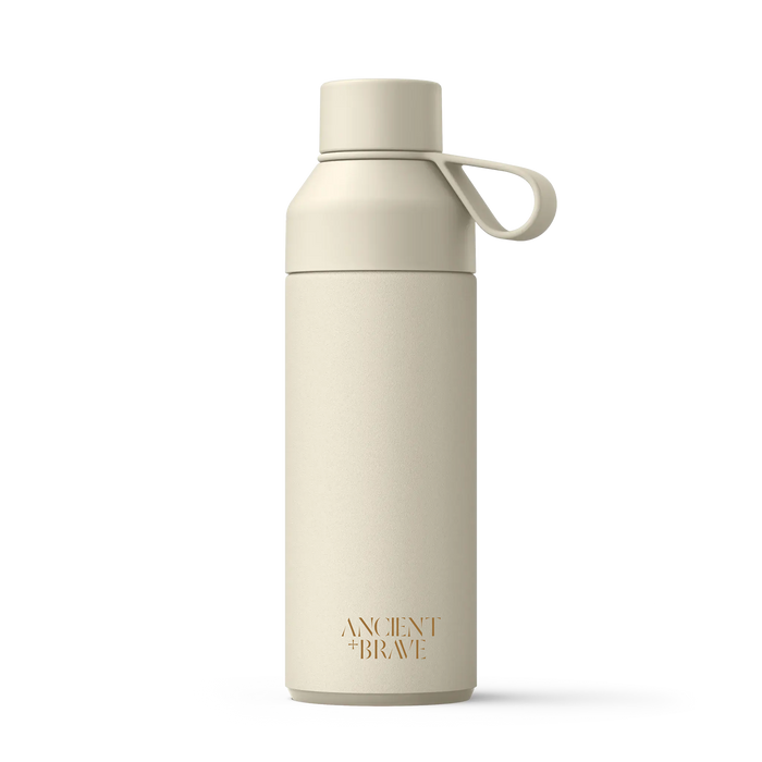 The Ancient + Brave x Ocean Bottle is a beige reusable water bottle with a screw-on lid and a carrying loop. Crafted from recycled stainless steel for an eco-friendly touch, it features the Ancient + Brave brand name in gold text near the base. The product is set against a plain white background.