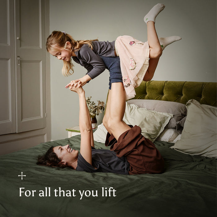 Woman lying on a bed with a child playfully lifting her leg, with text 'For all that you lift' on a neutral background.