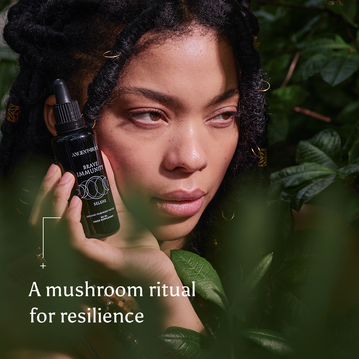 A person with long hair holds a black serum bottle labeled Brave Immunity from Ancient + Brave amid lush green leaves. Text on the image reads: A mushroom ritual for resilience, featuring functional mushrooms and organic botanicals to strengthen immunity.