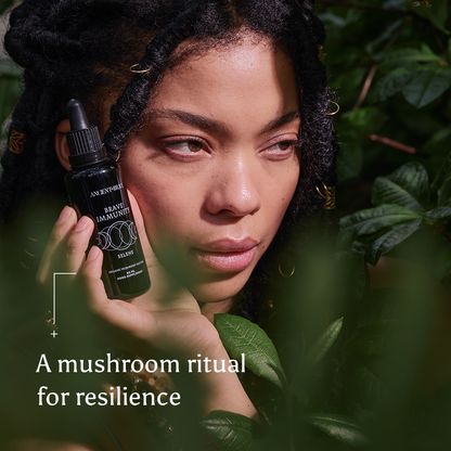 A person with long hair holds a black serum bottle labeled Brave Immunity from Ancient + Brave amid lush green leaves. Text on the image reads: A mushroom ritual for resilience, featuring functional mushrooms and organic botanicals to strengthen immunity.