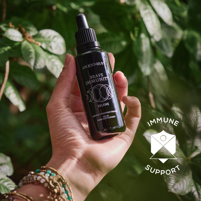 A hand holds a dark glass dropper bottle labeled “Brave Immunity” from Ancient + Brave, surrounded by lush greenery and organic botanicals. On the right side of the image, a white Immune Support icon and text overlay highlight the product’s potential to strengthen immunity.