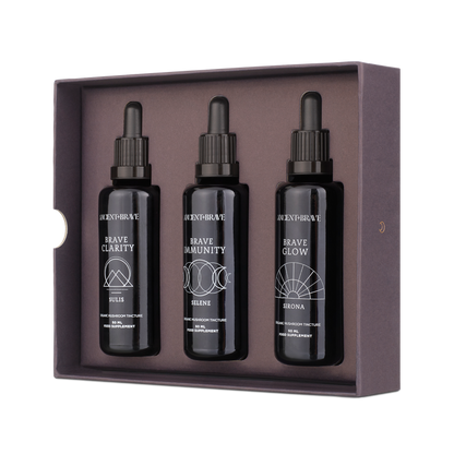 The Ancient + Brave Brave Tinctures Trio Set features three dark tinctures—Brave Clarity, Brave Immunity, and Brave Glow—elegantly arranged in a purple box adorned with white geometric patterns against a white background, making it an ideal choice to kickstart your wellness journey as inspired by the Ancient + Brave Journal.