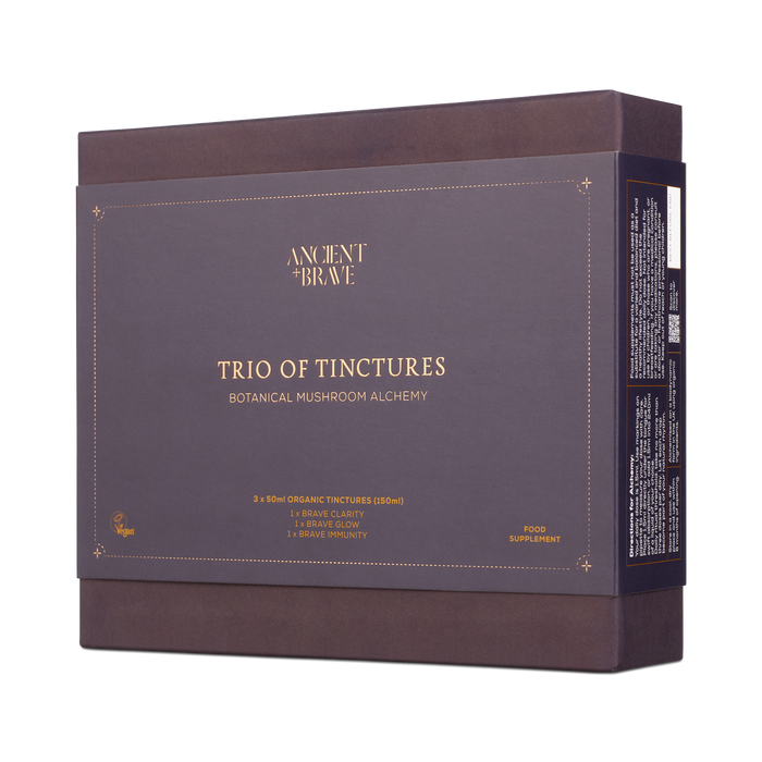 A dark purple box labeled Ancient + Brave—Brave Tinctures Trio Set, featuring product details, promotes clarity and is suitable for vegans. The set contains food supplements and includes insights from the Ancient + Brave Journal.