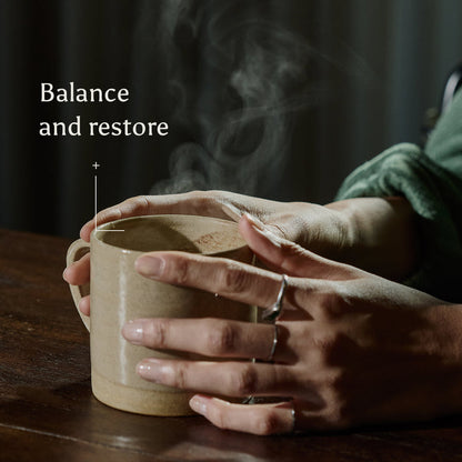 A person wearing rings holds a steaming mug of Ancient + Brave Cacao + Collagen with both hands on a wooden table. Text reads: Balance and restore. The soft, warm lighting invites you to nourish your skin from within.