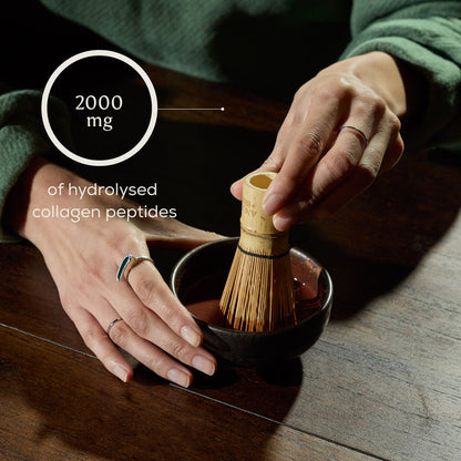 A person uses a bamboo whisk in a small bowl on a wooden table, blending Ancient + Brave Cacao + Collagen. Overlay text reads 2000 mg of hydrolysed bovine collagen peptides. Wearing a green sleeve and several rings, the individual creates a nourishing mix designed to support your skin from within.
