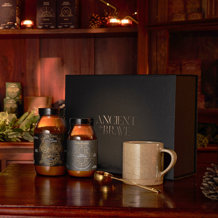 Displayed on a wooden table with dark wooden shelves and greenery in the background, the Ancient + Brave Davina Ritual Gift Set—including two jars, a mug, a gold spoon, and a black True Collagen box—showcases the benefits of hydrolysed peptides for optimum muscle function.