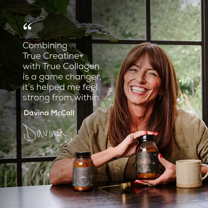 A smiling woman sits at a table holding a jar from the Ancient + Brave Davina Ritual Gift Set, with another jar, a spoon, and a quote about collagen’s support for muscle function beside her. Large windows and greenery are in the background.