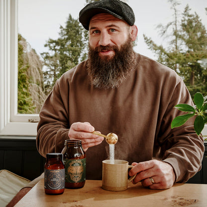 Joe Marler pouring a golden liquid from a spoon into a mug, with bottles on a table.
