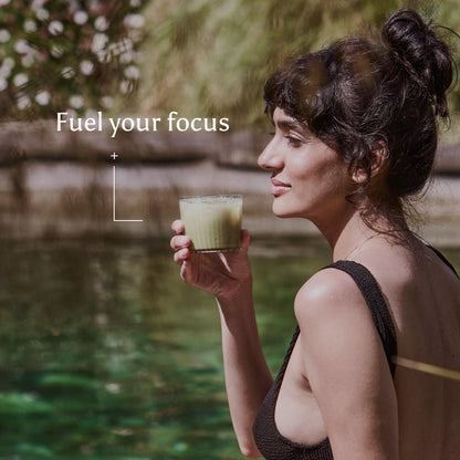 A woman in a black swimsuit sits by a pool, holding a glass with a light green drink made from Ancient + Brave Matcha + Collagen. White text in the top left corner reads Fuel your focus. Plants and water can be seen in the background.