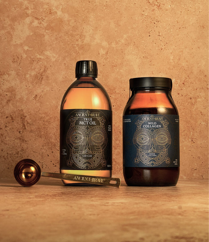 Two Ancient + Brave products from The Beginner’s Kit—a dark jar of Wild Collagen powder and a clear bottle of True MCT Oil—stand on a tan surface beside a gold measuring spoon. Each features an intricate dark blue and gold label, highlighting The Beginner’s Kit’s support for skin hydration and daily wellness.
