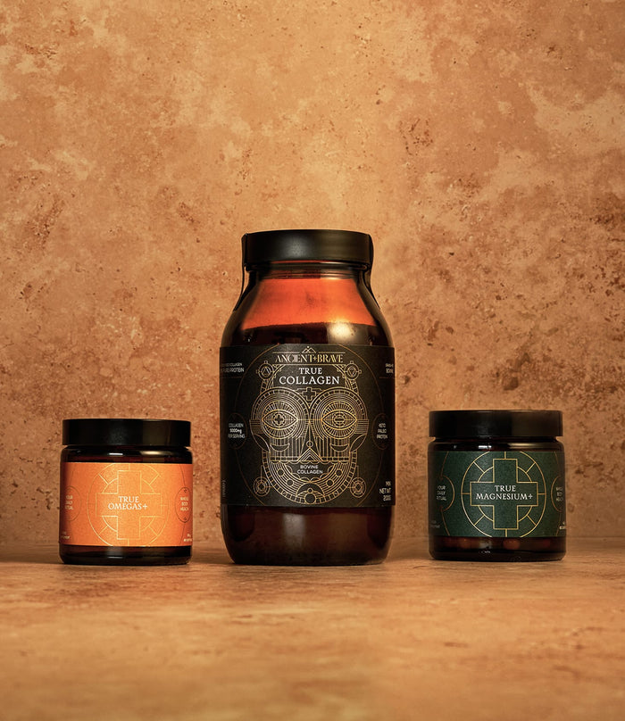 Three supplement jars from Ancient + Brave’s The Foundation Kit are displayed on a brown stone surface: one large jar in the center and two smaller jars on either side, labeled The Omegas and The Magnesium. This core supplement kit includes essentials like omega fatty acids to support your daily wellness routine.