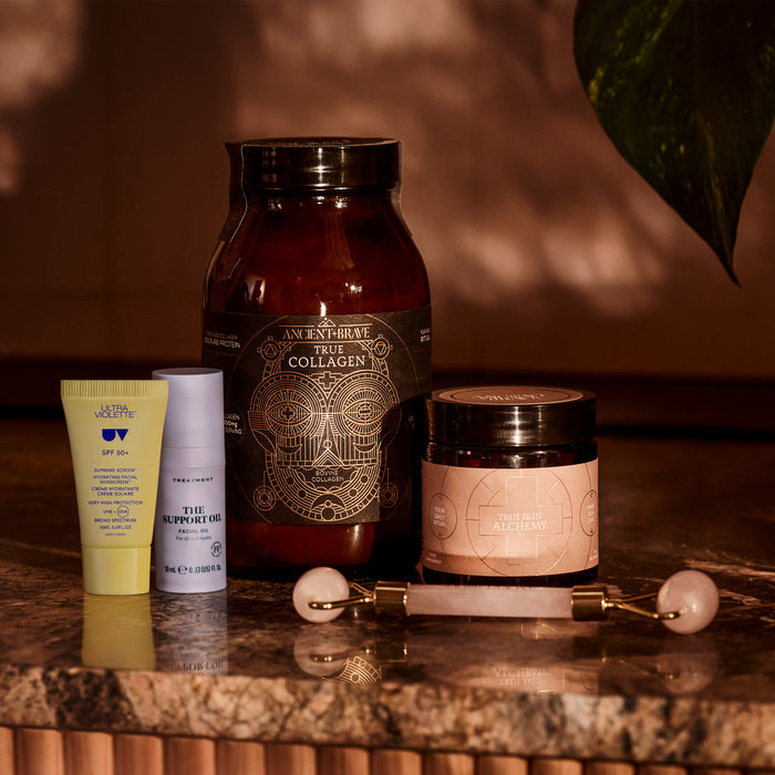Four skincare and wellness products from Ancient + Brave’s The Skin Essentials Kit—including a collagen jar, an oil bottle, a daily SPF sunscreen tube, a supplement jar for ingestible skincare benefits, and a pink facial roller—are arranged on a wooden surface with leafy shadows and warm lighting.