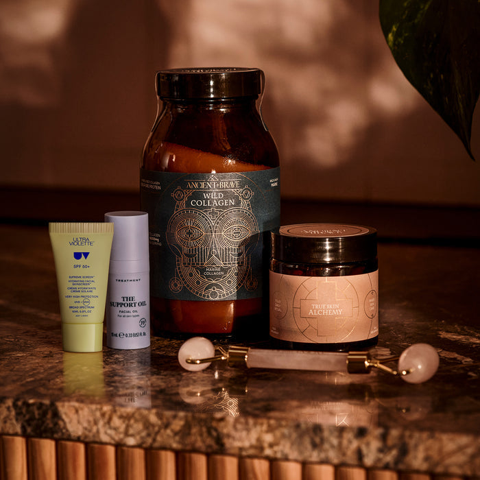 The Skin Essentials Kit by Ancient + Brave, featuring four expertly curated skincare products and a facial roller, is displayed atop a marble counter. Jars, a tube, and a bottle are thoughtfully arranged in warm lighting to evoke radiant-looking skin, with the soft presence of leafy greenery in the background enhancing the inviting scene.