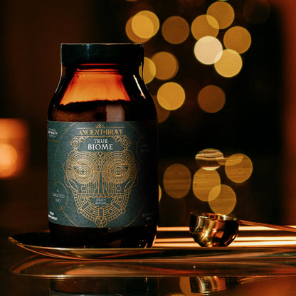 A brown jar labeled True Biome - Gift Subscription by Ancient + Brave is displayed on a reflective surface next to a gold measuring spoon, set against warm golden bokeh lights—an inviting scene that perfectly embodies wellbeing, making it an ideal choice for Christmas gift subscriptions.