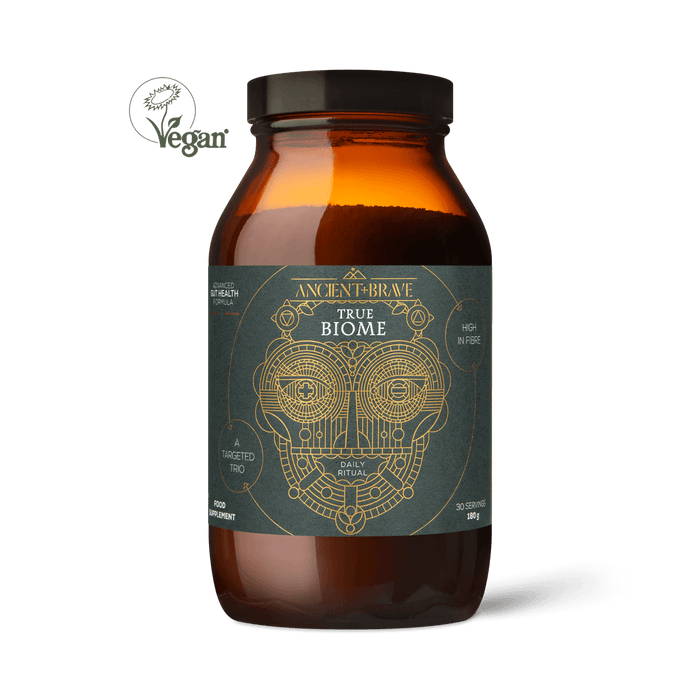 A brown glass jar labeled True Biome by Ancient + Brave, featuring a geometric face design, vegan symbol, and claims of high prebiotic fiber to support the gut barrier. The jar is displayed on a plain white background.