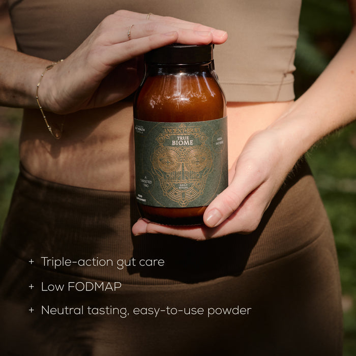 Person holding a bottle labeled 'True Biome' with text about product features including 'triple-action gut care, low FODMAP,  neutral tasting and easy-to-use powder.