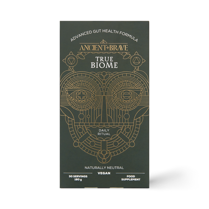 A sleek black box from Ancient + Brave, labeled “True Biome” and adorned with gold geometric patterns, features an advanced gut health formula containing prebiotics to support optimal gut barrier function. The packaging highlights it as a daily ritual, vegan-friendly, naturally neutral in flavor, and providing 30 servings.