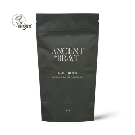 A dark charcoal gray stand-up pouch of Ancient + Brave True Biome supplement, featuring white and gold text, with a Vegan logo in the top-left corner on a white background.