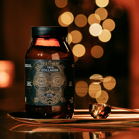 A jar of Ancient + Brave True Collagen - Gift Subscription powder sits on a reflective surface beside a metal scoop, with warm golden bokeh lights in the blurred background—ideal for boosting wellbeing.