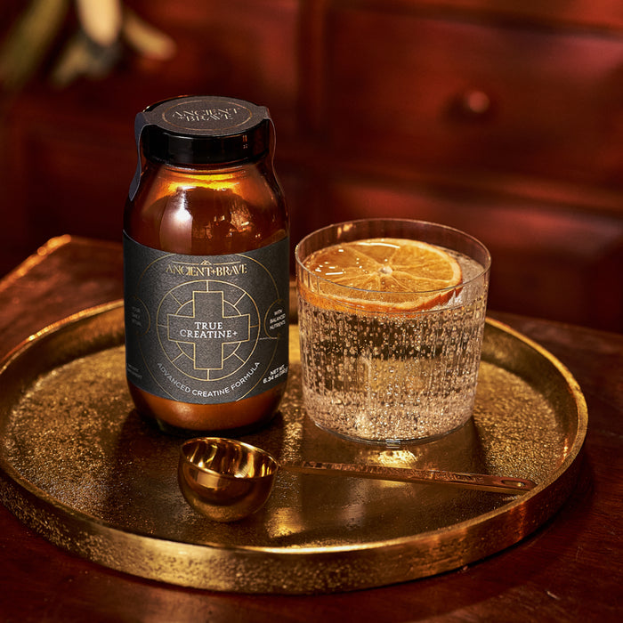 A jar of Ancient + Brave True Creatine+ - Gift Subscription rests on a gold tray beside sparkling water with dried orange and a gold spoon—an elegant wellbeing gift, all displayed on a wooden surface.
