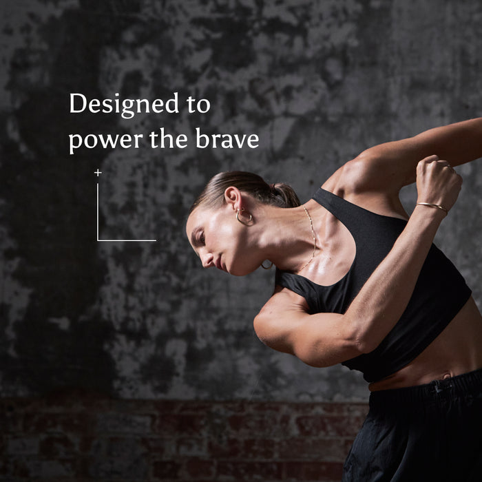 A strong woman in athletic wear leans sideways with one arm bent against a textured gray wall. Text reads, "True Creatine+ - Gift Subscription by Ancient + Brave: Designed to power the brave with wellbeing," with an intersecting lines graphic.