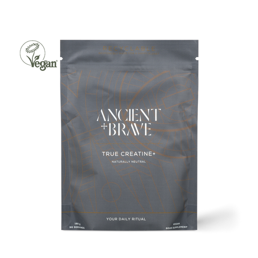 A dark gray resealable pouch labeled 'ANCIENT + BRAVE TRUE CREATINE+' with white text and thin orange line graphics, shown on a white background. A circular 'Vegan' logo appears to the left of the pouch.
