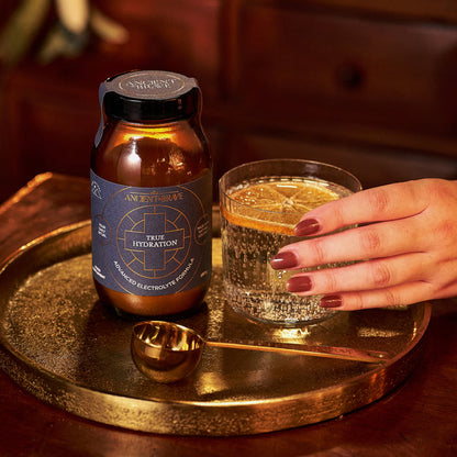A hand with painted nails holds a glass of sparkling water with lemon on a gold tray, capturing a vibrant moment of wellbeing. Beside it sits a jar of Ancient + Brave True Hydration and a gold spoon, subtly highlighting the True Hydration – Gift Subscription—a perfect choice for those who cherish true wellness rituals.