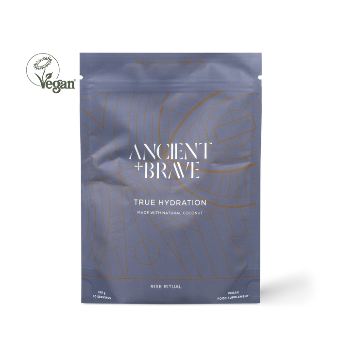 Ancient + Brawe True Hydration coconut powder supplement in a metallic slate blue pouch with gold line art, shown on a white background with a Vegan logo overlay.