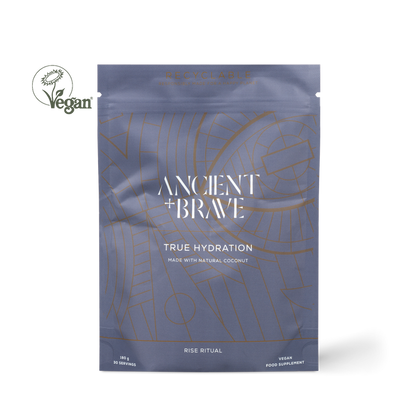 Ancient + Brawe True Hydration coconut powder supplement in a metallic slate blue pouch with gold line art, shown on a white background with a Vegan logo overlay.