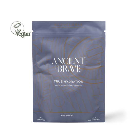 Ancient + Brawe True Hydration coconut powder supplement in a metallic slate blue pouch with gold line art, shown on a white background with a Vegan logo overlay.