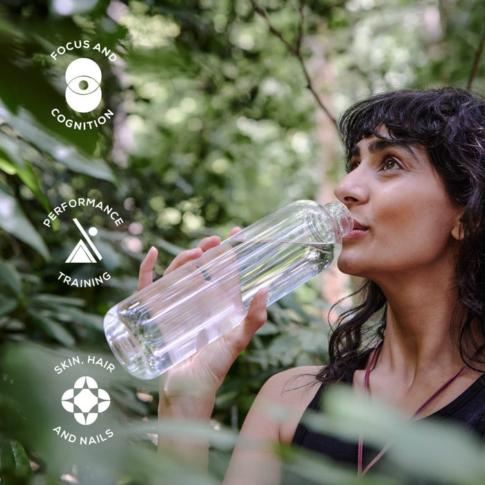 In a lush green forest, a woman drinks water enhanced with Ancient + Brave’s True Hydration - 3 Pack. Icons and text highlight its benefits: rapid rehydration with essential electrolytes to boost focus and cognition, support performance training, and promote healthier skin, hair, and nails.