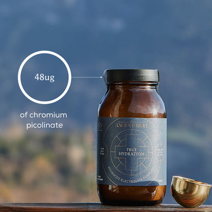 A brown glass jar labeled Ancient + Brave True Hydration is shown with a gold measuring scoop beside it. Text highlights 48μg of chromium picolinate and essential electrolytes for rapid rehydration. A blurred outdoor background with hills or mountains can be seen.
